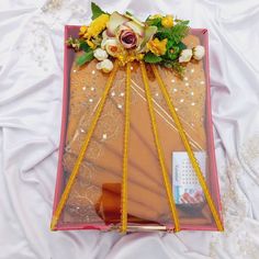 an arrangement of flowers and ribbons in a pink box on a white sheeted surface