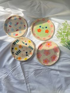 three plates with flowers painted on them sitting on a white sheet next to a plant