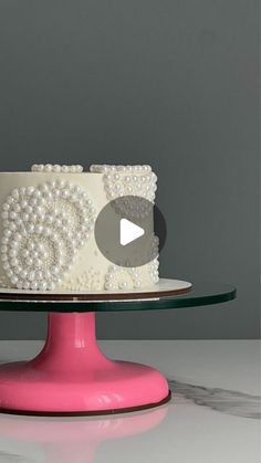 a white cake with pearls on it sitting on top of a pink plate next to a gray wall