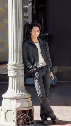 a young man leaning against a pole on the side of a street with his hands in his pockets