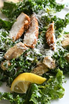 a white plate topped with greens and chicken covered in parmesan cheese next to a lemon wedge