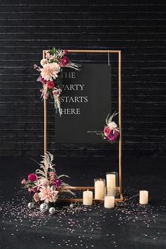 a sign that says the party starts here surrounded by candles and flowers on a black background