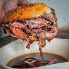 a hand is holding up a roast beef sandwich over a bowl of soup and dipping sauce