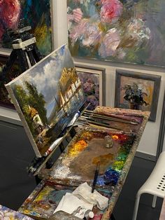 an artist's easel with paint and brushes on it in front of several paintings