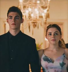 a young man and woman standing next to each other in a room with chandelier