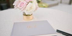 a white table topped with a vase filled with pink and white flowers next to a notebook