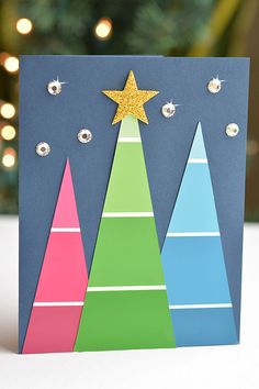 a handmade christmas card with three trees and a star on top, in front of a lit tree