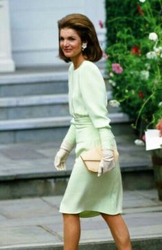 a woman in a green dress and white gloves walking down the street with flowers behind her