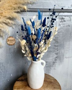 a white vase filled with blue and white flowers on top of a wooden table next to dry grass