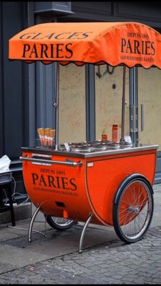 an orange cart sitting on the side of a street next to a table and umbrella