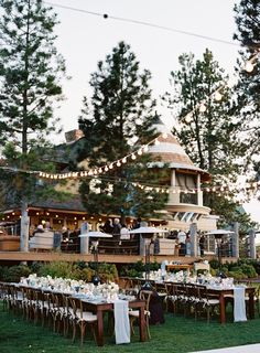 an outdoor dining area with long tables and white linens on the grass, surrounded by tall pine trees