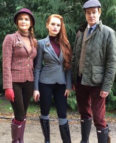 three people standing next to each other wearing well - worn boots and jackets with trees in the background