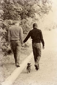 two men walking down a dirt road holding hands with a small dog in the foreground