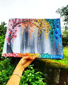 a person holding up a painting in front of some bushes and trees with colorful paint on it