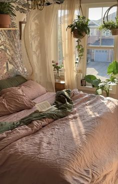 an unmade bed with pink sheets and pillows in front of a large window filled with potted plants