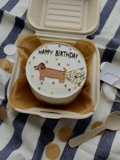 a birthday cake with a dog on it sitting in a box next to spoons