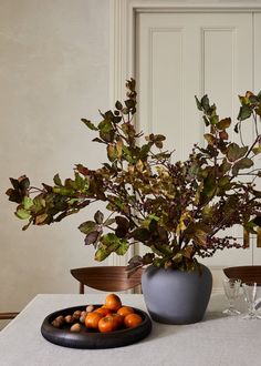 a table topped with a bowl of fruit next to a vase filled with leaves