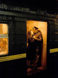 a man and woman standing on the door of a train at night, with their arms around each other