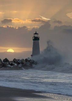 a light house on the beach with waves crashing in front of it and sun setting