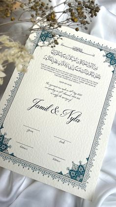 a wedding card with an ornate border on it and flowers in the background, sitting on a white sheet