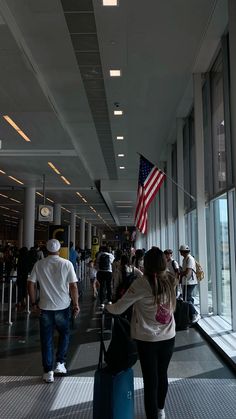 people walking through an airport with luggage and american flag hanging from the ceiling in front of them