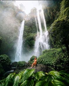 a woman sitting in front of a waterfall with lush green foliage on the ground and trees around her