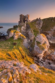 an old castle sits on the edge of a cliff overlooking the ocean at sunset or dawn