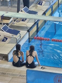 two women in bathing suits sitting on the edge of a swimming pool while looking at something