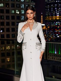 a woman standing in front of a window wearing a white dress and jacket with sequins on it