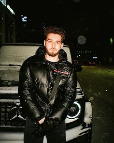 a man standing in front of a white car with his hands on his hips while wearing a black jacket