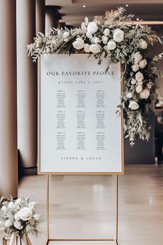 a sign with flowers and greenery on it in the middle of a room next to two vases