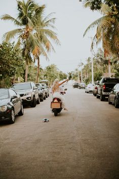 a woman riding on the back of a scooter down a street next to parked cars