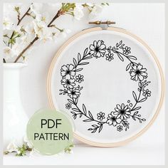 a white vase with flowers on it next to a cross stitch embroidery pattern and a floral wreath