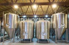 three stainless steel tanks in a warehouse with lights on the ceiling and windows above them