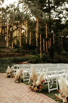 an outdoor ceremony setup with white chairs and pamodia flowers on the aisle, surrounded by string lights