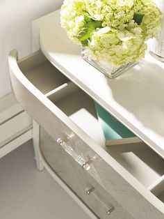 a vase with flowers sitting on top of a white table next to a mirror and drawer