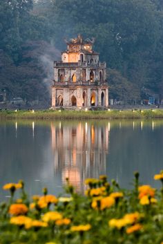 an old building sitting on top of a lake surrounded by trees and flowers in the foreground