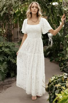 a woman wearing a white dress standing in front of trees and plants with her hand on the ground