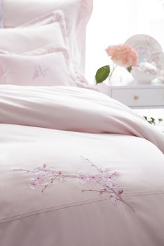 a bed with pink sheets and pillows in a room next to a flower vase on a dresser