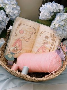 a basket filled with yarn next to blue flowers and an open book on top of a table