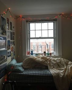 an unmade bed in front of a window with christmas lights strung across the room