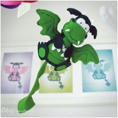 a green stuffed dragon hanging from the ceiling in front of three framed pictures on a wall