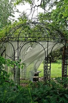 an outdoor gazebo in the middle of some bushes and trees with a white cloth draped over it