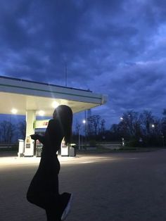 a woman standing in front of a gas station at night with her shadow on the ground