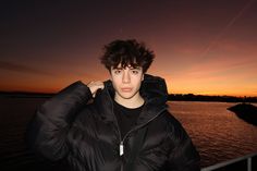 a young man is standing by the water at sunset with his jacket on, looking off into the distance