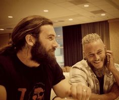 two men sitting next to each other at a table talking on their cell phones and smiling