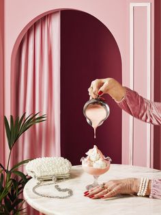 a woman pouring something into a bowl on top of a table with pink walls and curtains