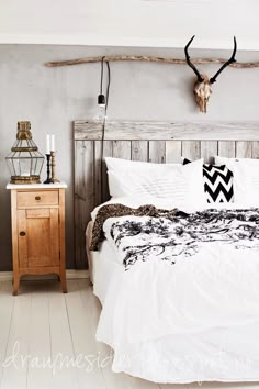 a bed with white sheets and pillows in a bedroom next to a wooden dresser, lamp and deer head