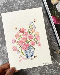 a hand holding a card with watercolor flowers in a blue and white vase on it