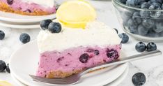 a slice of blueberry cheesecake on a plate with a fork and bowl of blueberries in the background